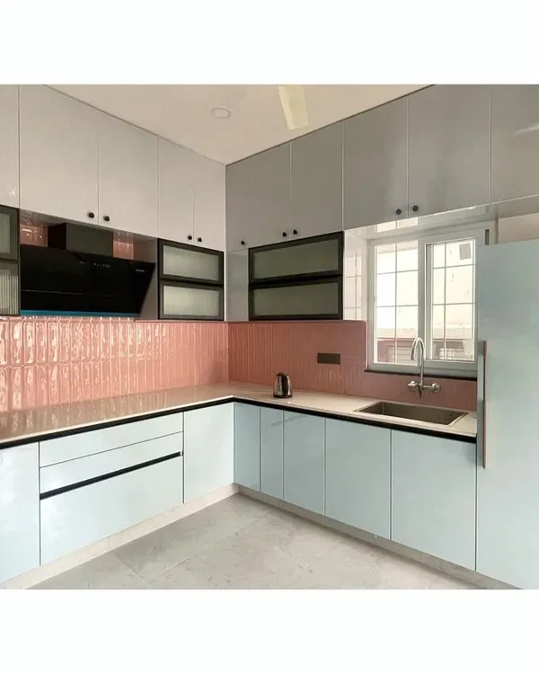 This L-shaped modular kitchen features a unique combination of glossy light blue base cabinets and a striking pink tiled backsplash. The black-framed glass upper cabinets add a modern, industrial touch to this vibrant and functional cooking space.
