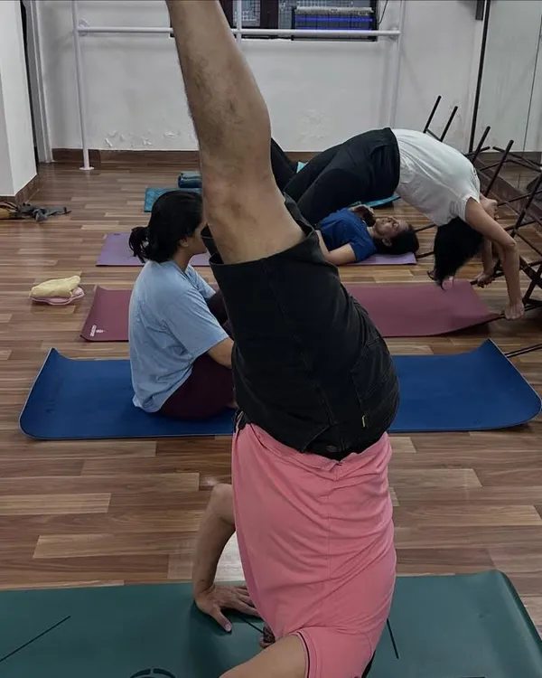 A student practices a headstand while another works on a supported backbend in the background, showing the diverse focus within a single class.