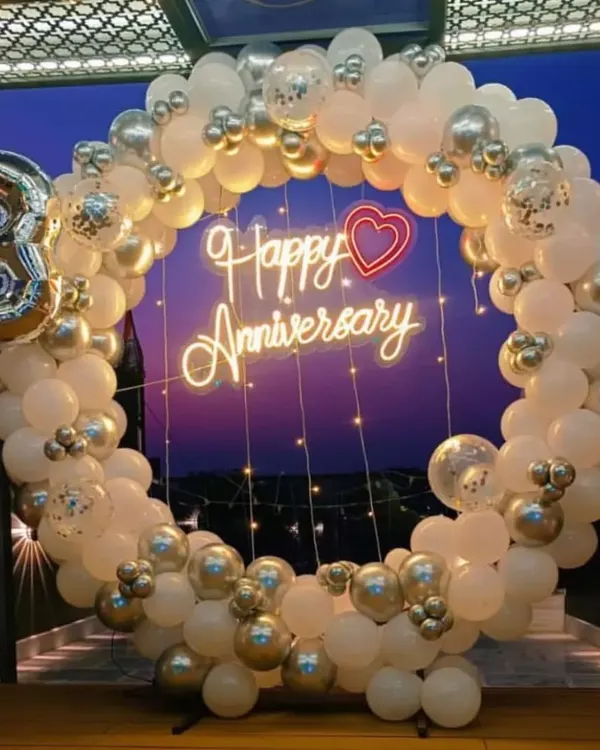 A beautiful anniversary backdrop featuring a full circle balloon arch in white and silver chrome. The design is completed with a "Happy Anniversary" neon sign and a large number "3" balloon for a third anniversary.
