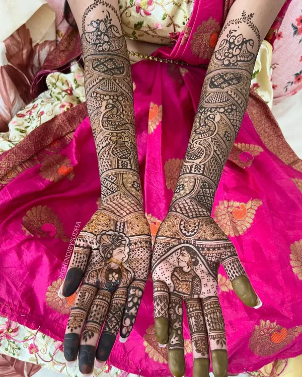 A complete bridal mehendi look featuring portraits of the bride and groom on the palms. The design extends up the forearms with traditional motifs like peacocks and kalash (pots), symbolizing prosperity and love for the new journey ahead.