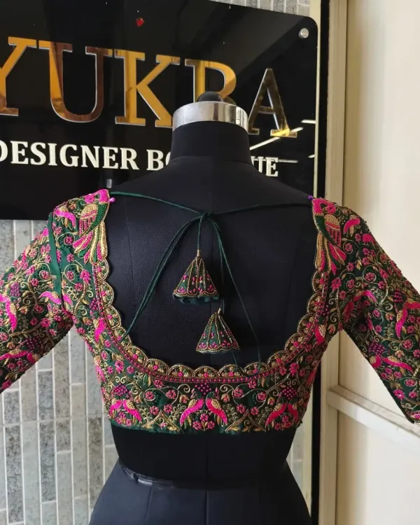 This emerald green blouse features a wide U-shaped back with a beautifully scalloped border. The entire back and sleeves are covered in dense pink and gold floral embroidery, making it a true work of art.