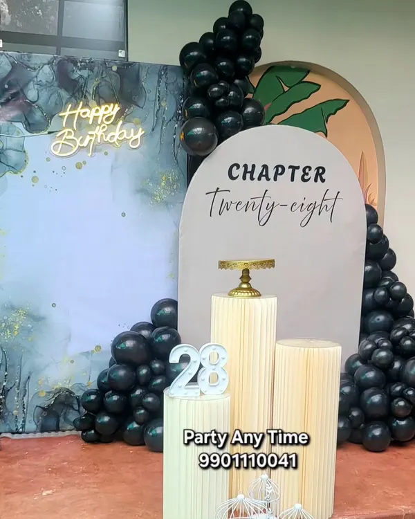 A sophisticated "Chapter Twenty-eight" birthday setup. This features a marble-effect backdrop, a modern arch panel, black balloons, and elegant white plinths.