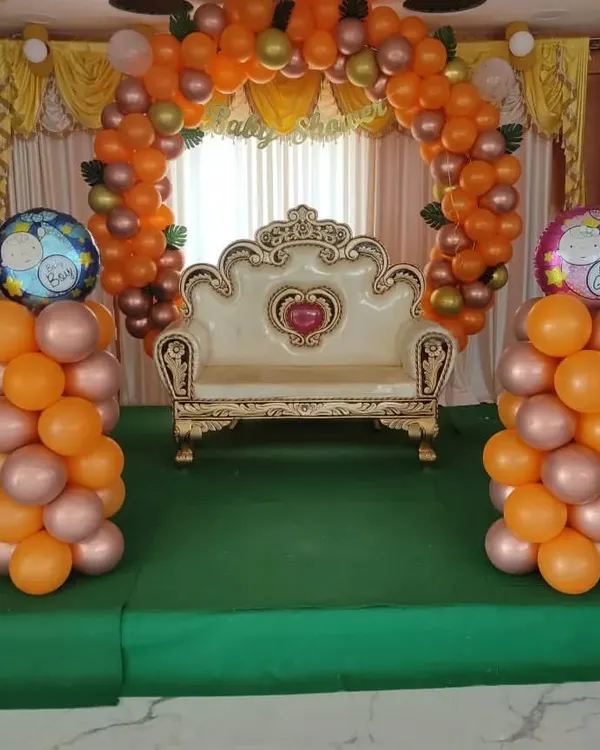 A traditional baby shower stage setup with an orange balloon arch and a decorative sofa for the mother-to-be.