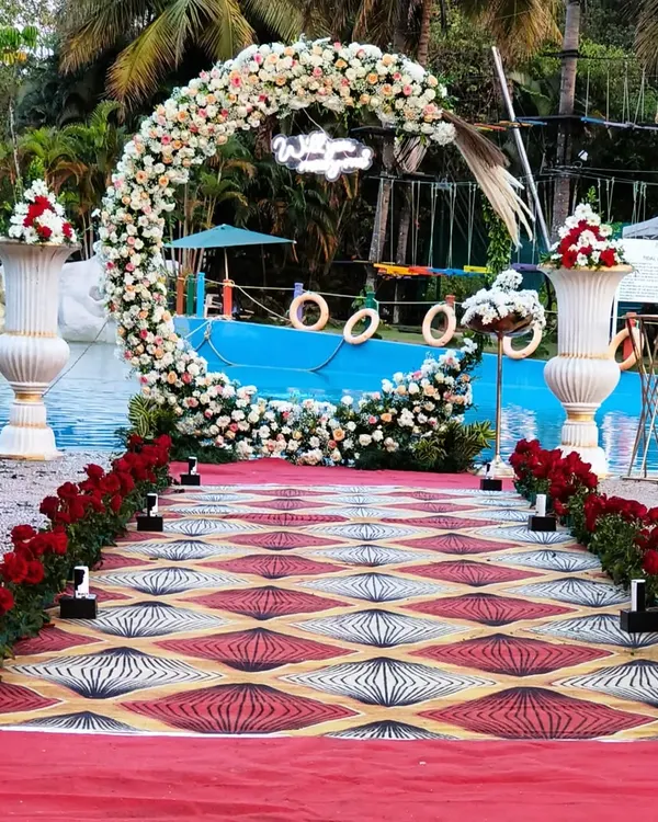 This poolside entrance features a circular floral arch with a "Welcome" sign, leading guests down a custom-printed aisle runner. The red rose border adds a classic touch to this modern outdoor wedding setup.