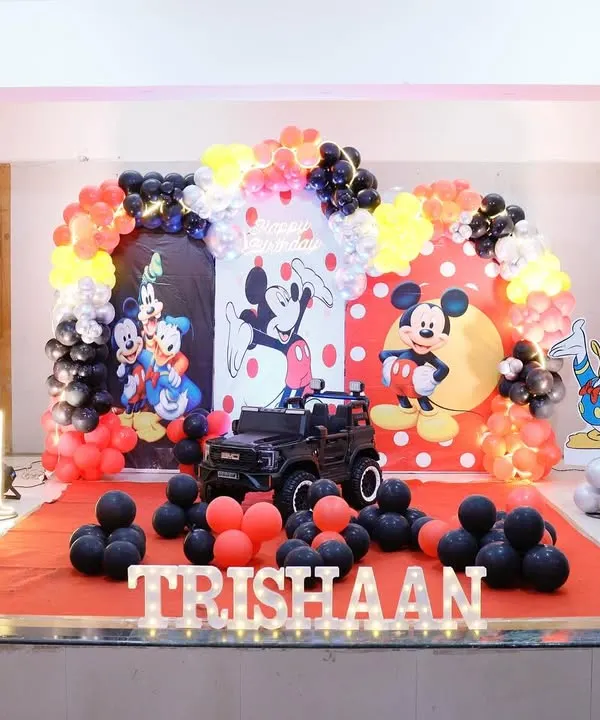 A grand Mickey Mouse themed first birthday party setup. We created a full stage with a vibrant red, yellow, and black balloon arch, custom backdrops, character cutouts of Mickey, Donald, and Pluto, and personalized light-up letters for the birthday boy.