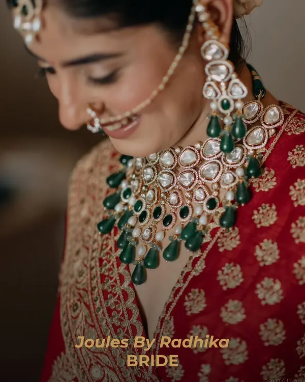 Not just a bride, but a vision. This close-up captures the radiant smile of a JBR Bride, adorned in a bespoke polki necklace with cascading emerald drops, perfectly complementing her red bridal attire.
