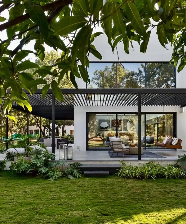 The exterior of the Mango Orchard House shows how the structure opens up to the landscape. A large, shaded terrace extends the living space into the garden, creating a perfect setting for outdoor living while being sheltered by a modern pergola.