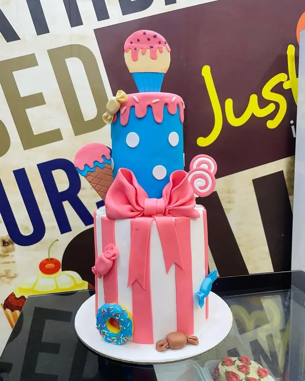 A playful two-tier custom cake designed for a fun-filled party. The bottom tier features pink and white stripes with a large fondant bow, while the top blue polka-dotted tier is topped with a melting ice cream cone, lollipops, and other candy decorations.