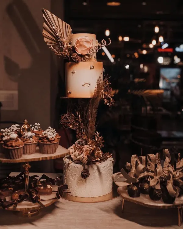 An aesthetic engagement dessert table with a rustic, bohemian theme. The setup includes a two-tier cake, matching cupcakes, and other small desserts, all decorated with dried flowers and pampas grass.