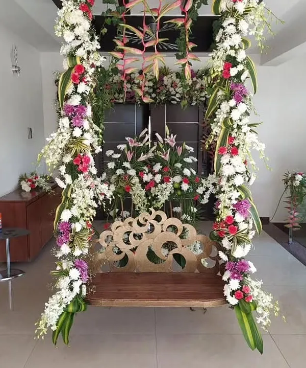 A beautifully decorated traditional wooden swing (jhoola) for a housewarming or pre-wedding ceremony at home. The swing is heavily adorned with vertical garlands of white, red, and purple flowers and exotic leaves.