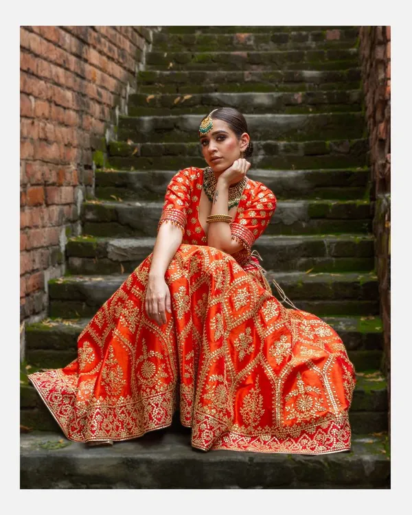A full-length portrait of a model in a vibrant orange and gold embroidered lehenga, seated on rustic stone steps. This shot highlights my ability to capture the grandeur of Indian bridal couture in an outdoor, editorial setting.