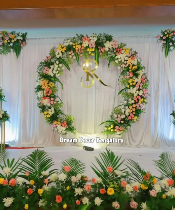 A beautiful and classic engagement decor. The large floral ring with the couple's initials, set against a simple white drape, creates an elegant and personalized stage.