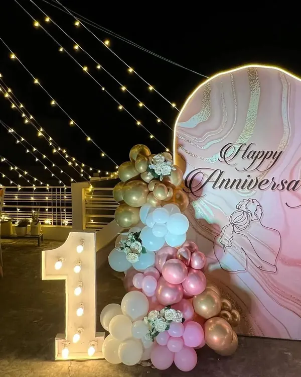 A beautiful first anniversary celebration on a terrace under string lights. The setup features a custom arched backdrop with a marble texture, a balloon arrangement, and a large light-up number '1'.