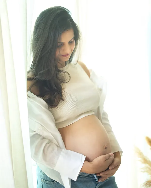 A serene and beautiful solo portrait. The soft window light illuminates the mom-to-be, who is dressed in a casual crop top and jeans, creating a gentle and authentic image.
