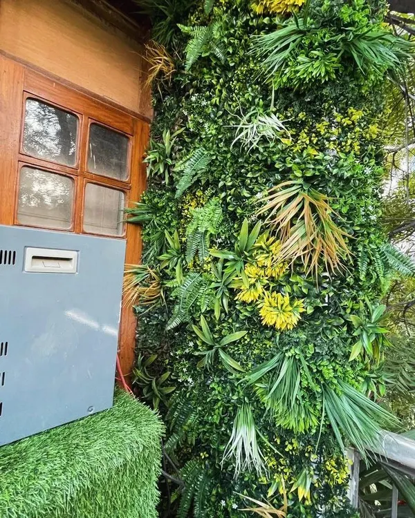 A dense and textured artificial green wall installed next to a wooden window frame. The variety of faux ferns, leaves, and yellow accents creates a realistic, jungle-like effect.