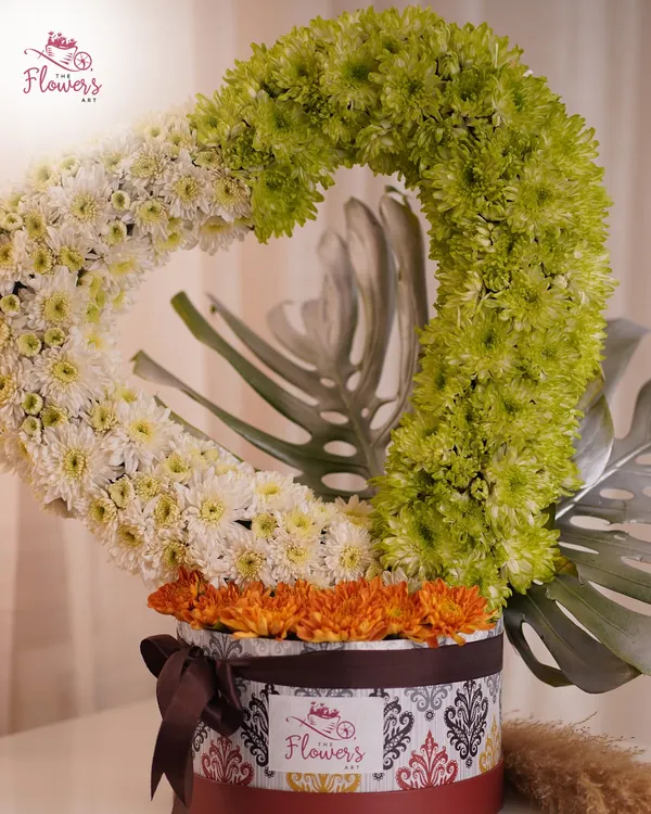 A modern and artistic floral sculpture featuring a heart shape made from densely packed green and white chrysanthemums. This unique design sits atop a decorative box filled with orange daisies.