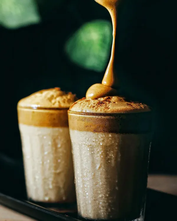 Capturing the perfect pour of creamy Dalgona coffee into a glass. The dark, moody background with soft green highlights makes the texture of the foam the hero of this shot.