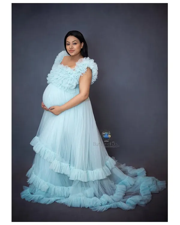A stunning solo maternity portrait showcasing the mother-to-be in a flowing, light blue ruffled gown against a classic studio backdrop.