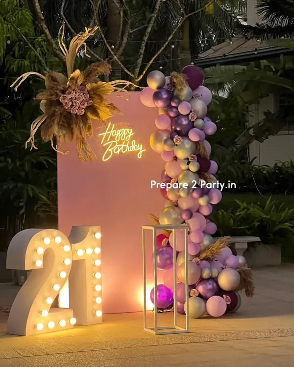 A chic and modern 21st birthday setup in an outdoor garden setting. This design features large light-up marquee numbers, a stylish pink backdrop, and a beautiful balloon garland with pampas grass and floral details.