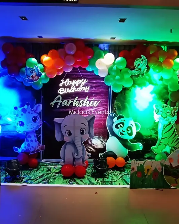 A wide view of the complete jungle safari theme setup for Aarshiv's birthday. The decor features a full balloon arch, a personalized neon sign, and multiple cute animal cutouts like a lion, giraffe, elephant, and panda.