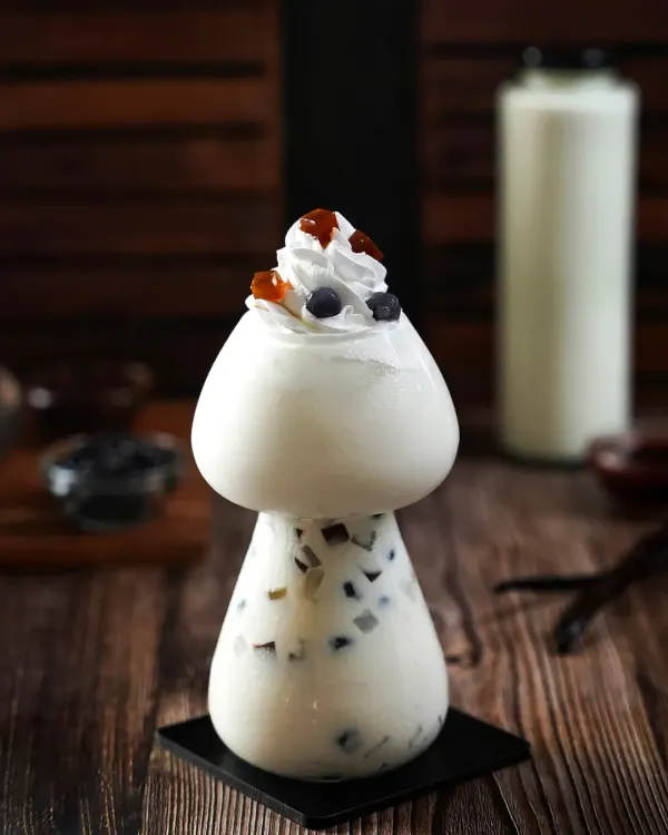 A whimsical mushroom-shaped glass filled with a creamy boba drink. The dark, woody background and moody lighting create a magical, forest-like atmosphere that makes the drink feel unique and enchanting.