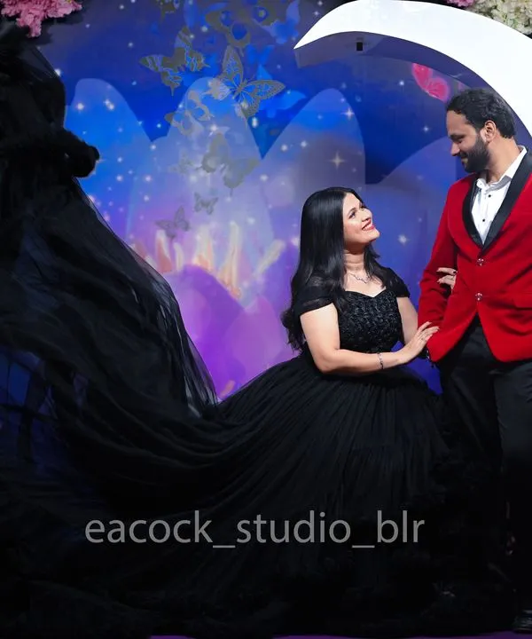 A dramatic pre-wedding studio shot featuring a flowing black trail gown and a bold red blazer. The moon prop and floral background create a fantasy-like, romantic atmosphere.