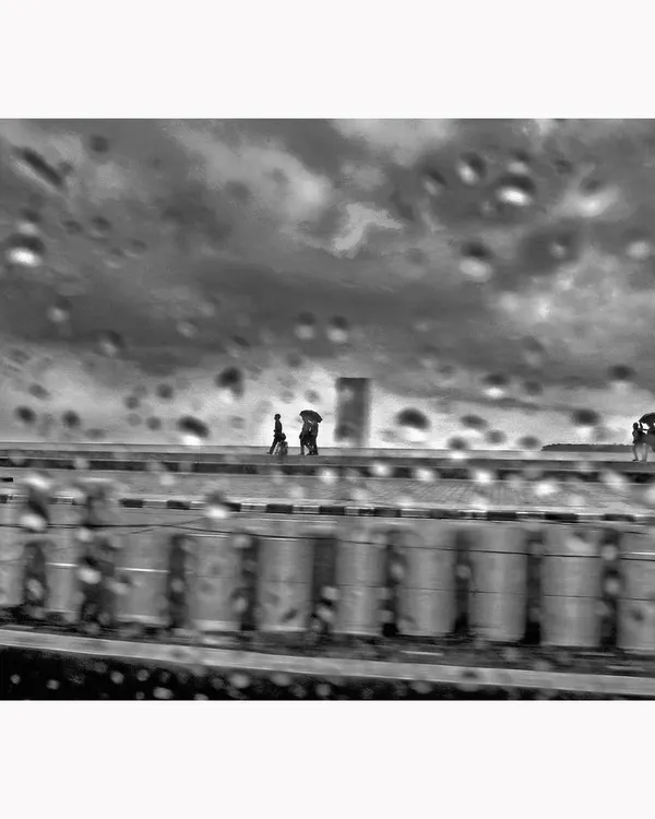 A moody, atmospheric scene of a rainy day, viewed through a water-streaked window. The motion blur and droplets create a painterly effect, turning a mundane moment into a piece of evocative black and white street photography.