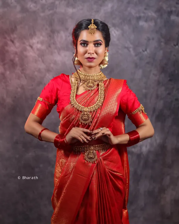 A full-length studio portrait of a model in a stunning red Kanjivaram silk saree. The focus is on the complete bridal look, from the temple jewelry and waist belt (vaddanam) to the traditional drape of the fabric.