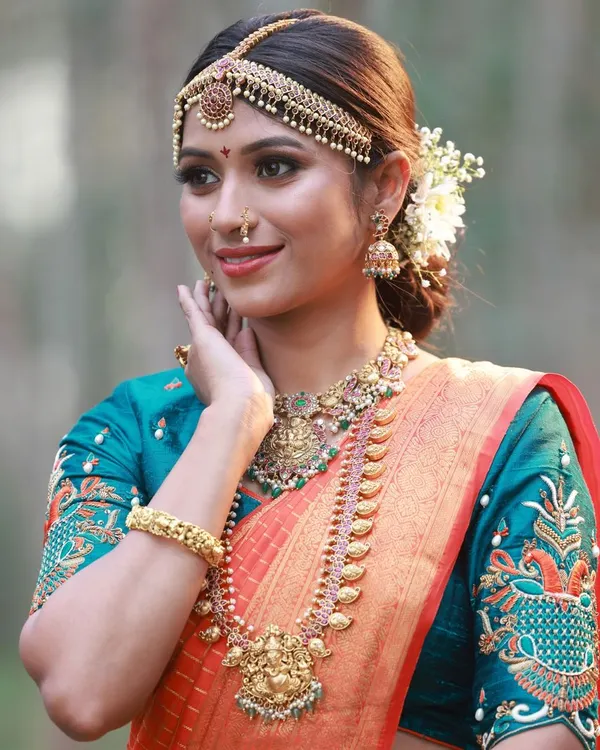 A beautiful close-up of a South Indian bridal look. The focus is on the detailed makeup, the traditional temple jewellery, and the rich colors of the orange and teal silk saree.