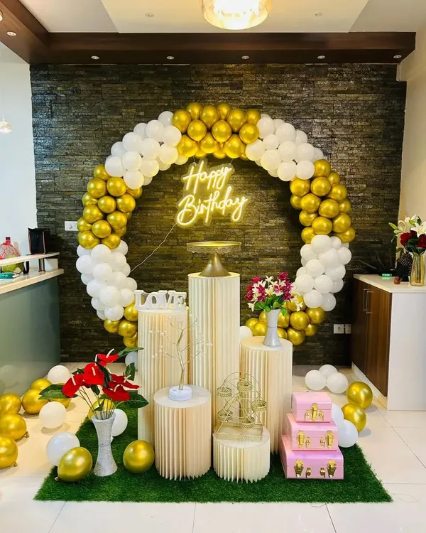 A classic and elegant birthday setup with a full circular balloon arch in gold and white. The decoration is placed on an artificial grass mat and includes pedestals, floral arrangements, and a 'Happy Birthday' neon sign.