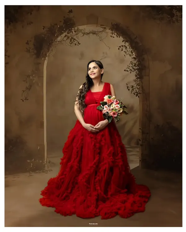 A full-length portrait of a mother-to-be in a stunning red ruffled gown, holding a bouquet of flowers against a rustic archway backdrop. This is a perfect example of our fine art maternity portraiture.