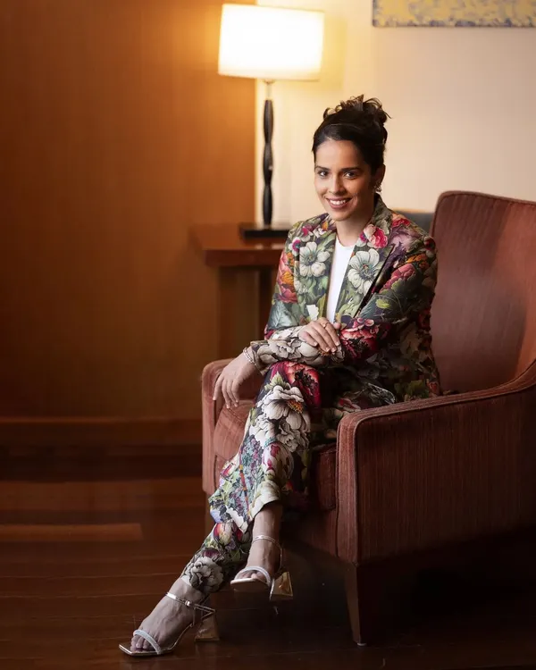 A full-length portrait of Saina Nehwal in a stylish floral pantsuit. The relaxed, seated pose in a well-lit room creates a look that is both professional and effortlessly chic.