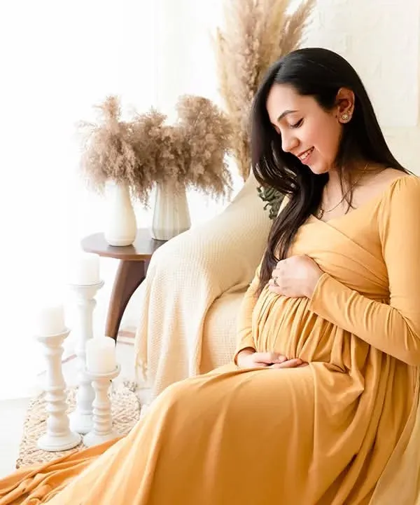 A solo maternity portrait in a mustard yellow gown, set against a bright, boho-chic studio setup. The natural light and earthy decor elements create a warm and inviting atmosphere.