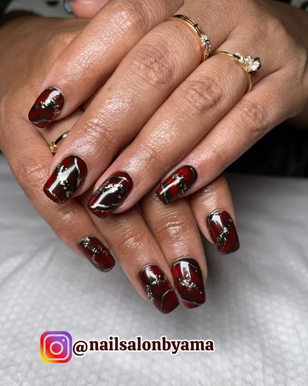 A stunning close-up of our custom nail extensions. This design features a rich, deep red and black marble effect, highlighted with elegant gold foil accents for a luxurious and bold statement.