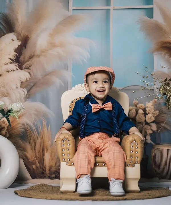 A dapper young boy in a bow tie and cap smiles from his miniature armchair. This studio portrait uses a beautiful boho-themed backdrop with pampas grass for a stylish and warm feel.