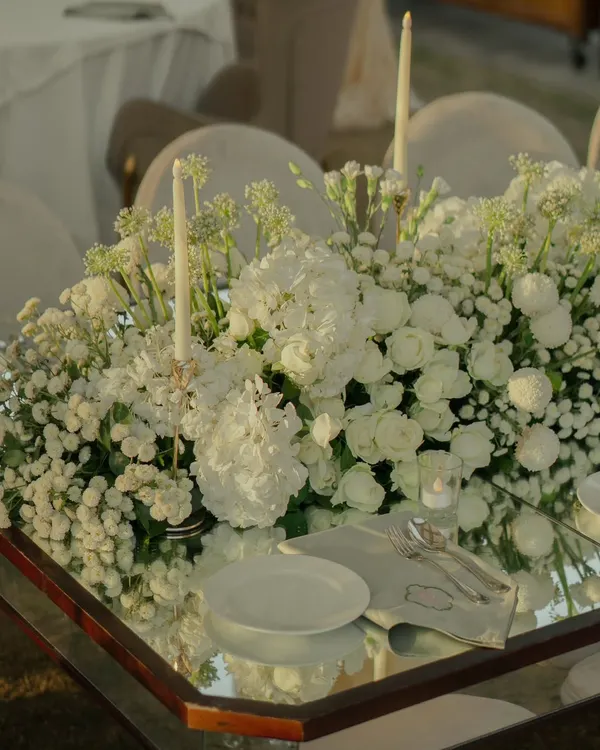 A mirror-top table reflects a lush arrangement of white hydrangeas, roses, and baby's breath, paired with elegant taper candles for a timeless and romantic look.