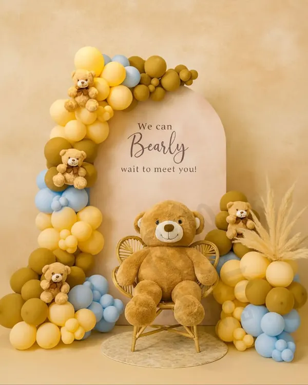 The classic 'We can Bearly wait' baby shower theme. This setup features a charming balloon arch in pastel yellow, blue, and brown, with small teddy bears nestled in the balloons and a large one seated on a wicker chair.