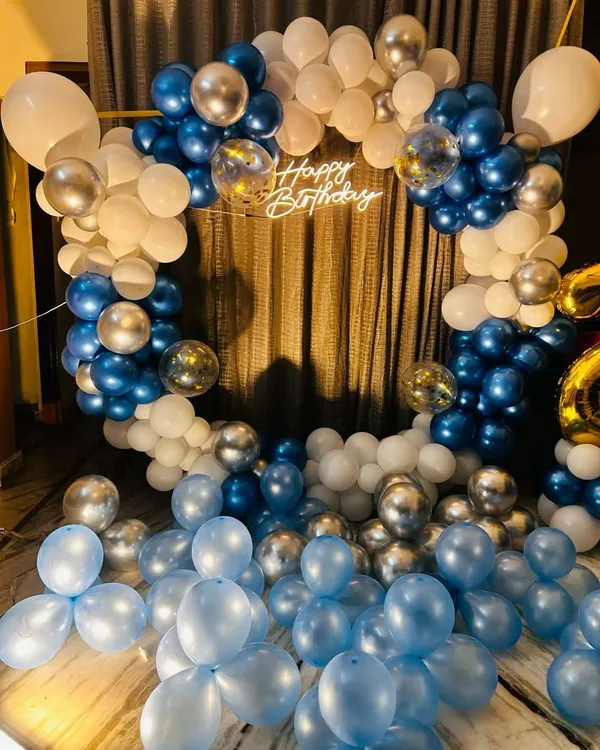 A classic home birthday decoration featuring a full ring balloon arch in blue, white, and silver chrome. The 'Happy Birthday' sign in the center and balloons scattered on the floor create a festive and immersive feel.