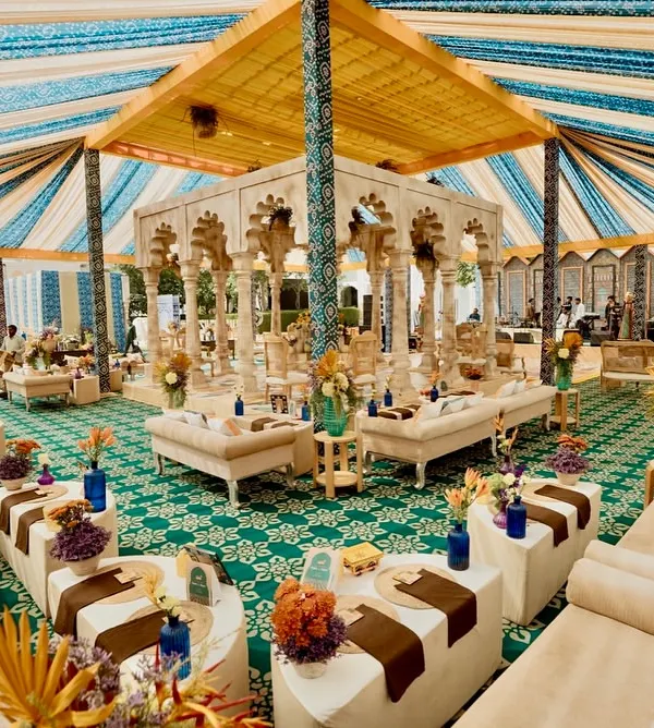 A wide view of the 'Rang Mahal' welcome lunch, inspired by Rajasthani elegance. A central pavilion with carved stone columns is surrounded by plush lounge seating, all under a vibrant blue and yellow canopy.