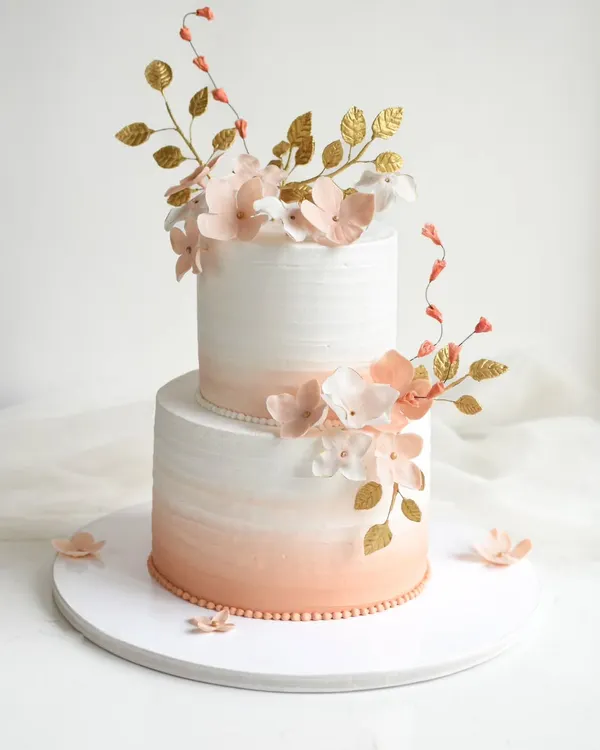 This two-tiered cake is a perfect example of soft elegance, featuring a delicate peach ombre effect in white chocolate ganache. It's adorned with handcrafted sugar flowers and touches of gold leaf, making it a dreamy showstopper for weddings or engagements.