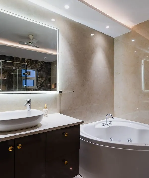This bathroom design blends elegance and modernity. It features a corner jacuzzi tub, a sleek vanity with a vessel sink, and an illuminated mirror, all set against warm beige marble tiles.
