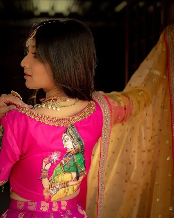 This bright pink raw silk blouse features a beautiful, hand-painted depiction of a traditional figure, making it a wearable piece of art. The delicate gold embroidery along the neckline adds a touch of festive elegance.