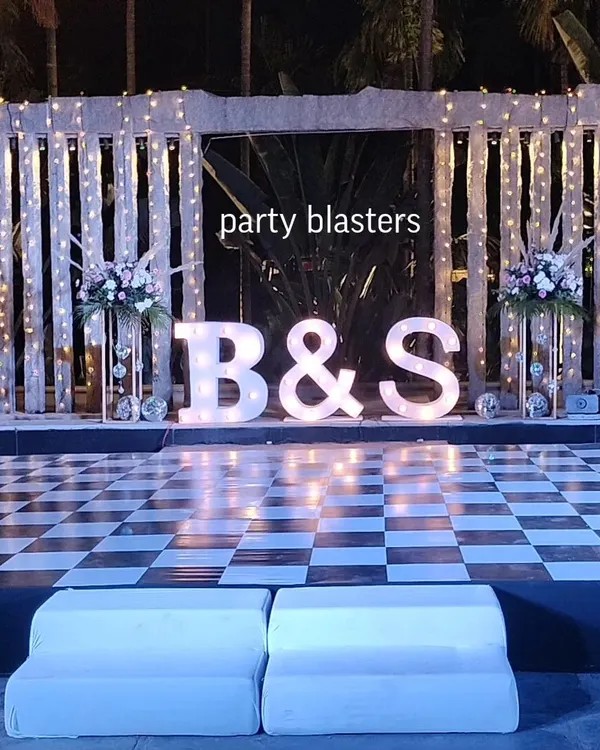 A high-energy Sangeet stage ready for a night of dancing. The large, illuminated marquee letters with the couple's initials, set against a lit backdrop and a checkered dance floor, create a fun and personalized party atmosphere.