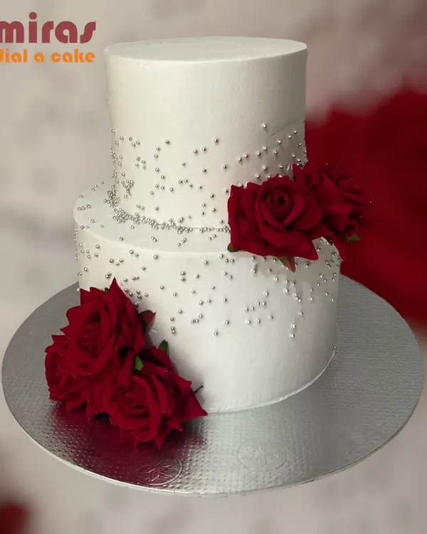 This elegant two-tier cake features a clean white buttercream finish, delicate silver beadwork, and deep red roses. It's a sophisticated choice for a formal corporate anniversary or celebration.