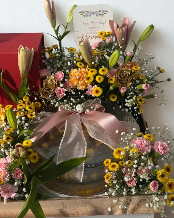 A beautiful birthday hamper where gifts and fresh flowers come together to create a stunning display. This arrangement features a vibrant mix of lilies and carnations alongside carefully selected presents, all tied together with delicate pink ribbons.