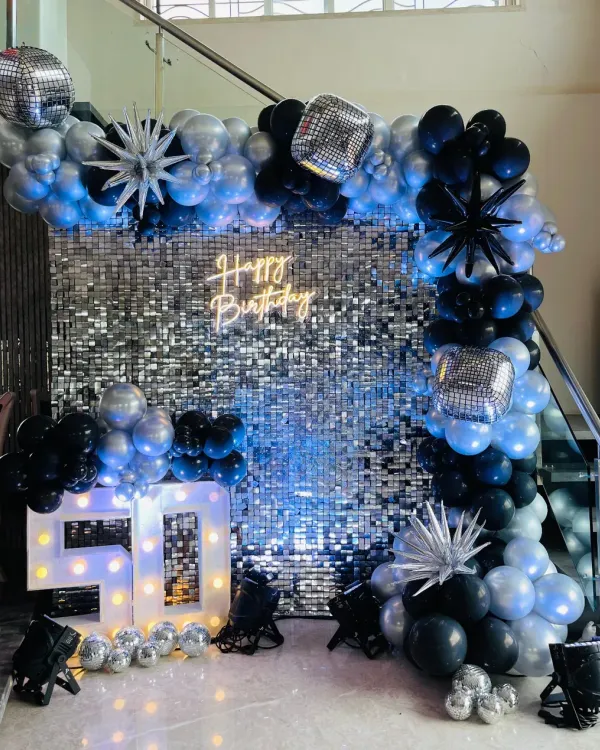 A glamorous 50th birthday setup featuring a full silver sequin wall, a "Happy Birthday" neon sign, and large light-up marquee numbers. The black and silver balloon arch is accented with disco balls and starburst props for a complete party look.