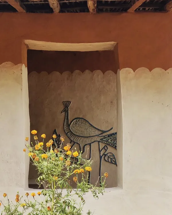 A detail from a vernacular homestay in Madhya Pradesh. The walls are finished with mud and decorated with a traditional peacock mural, framed by a pot of marigolds. This is architecture that celebrates local art and materials.