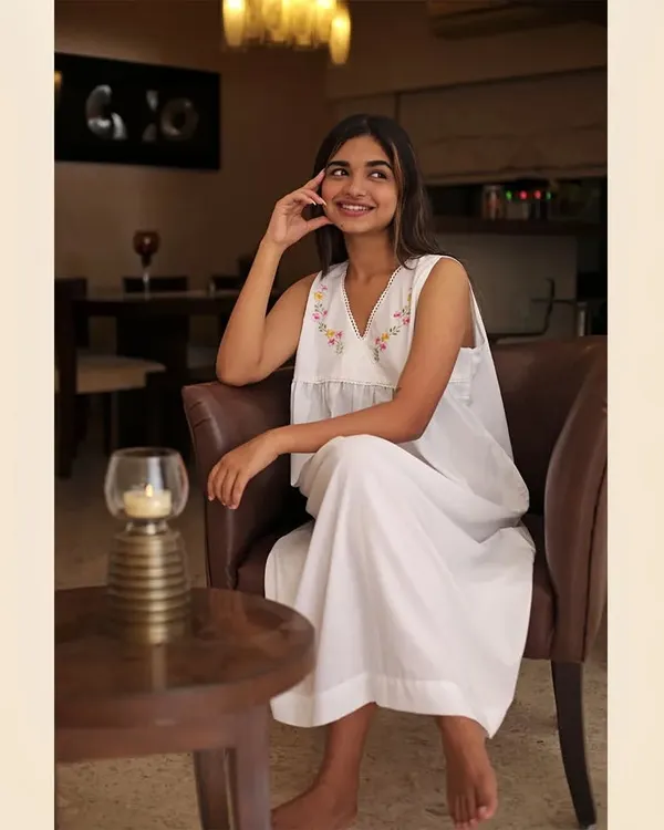 A lifestyle shot for a loungewear brand, set in a warm, ambient living room. The model's relaxed pose and smile convey comfort, making this white embroidered set look perfect for a quiet evening at home.