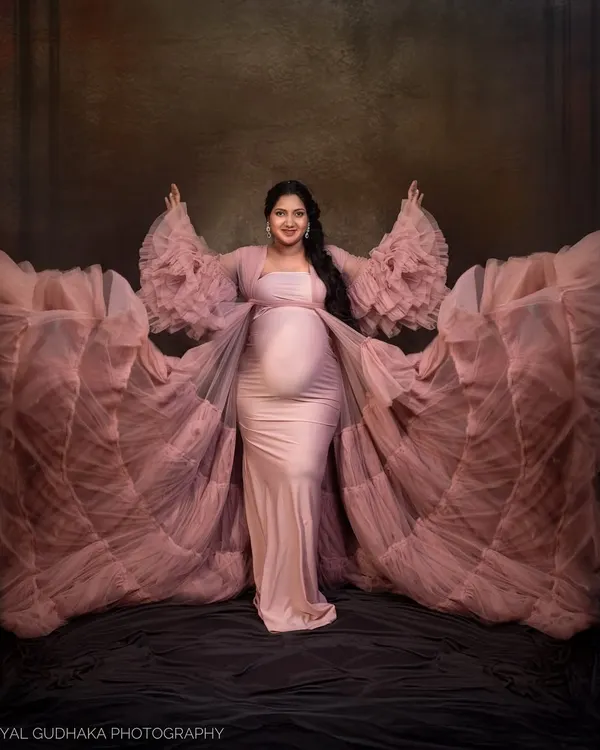 Embracing the power and beauty of motherhood. This mom-to-be shows off our popular pink ruffled gown, its dramatic sleeves creating a stunning, angelic silhouette against a classic painterly backdrop.