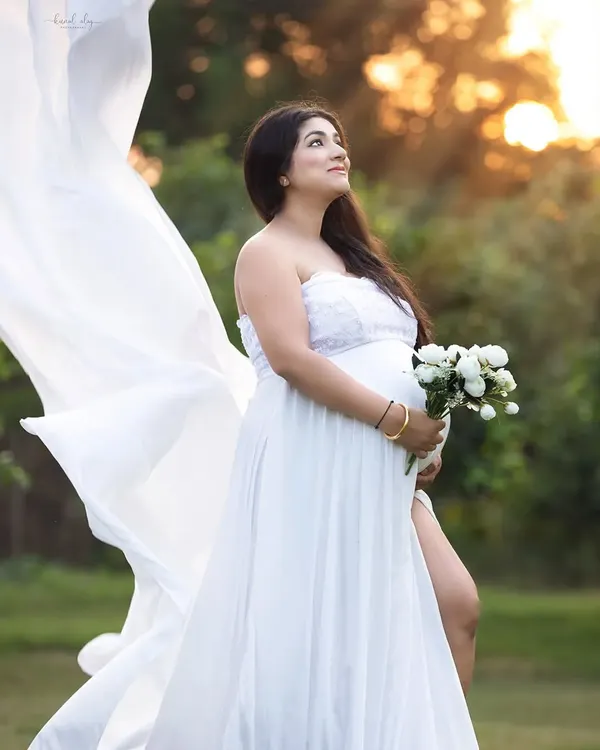 An outdoor maternity portrait taken during the golden hour, with soft sunlight creating a warm, ethereal glow. The flowing white gown and natural setting provide a romantic and serene feel.
